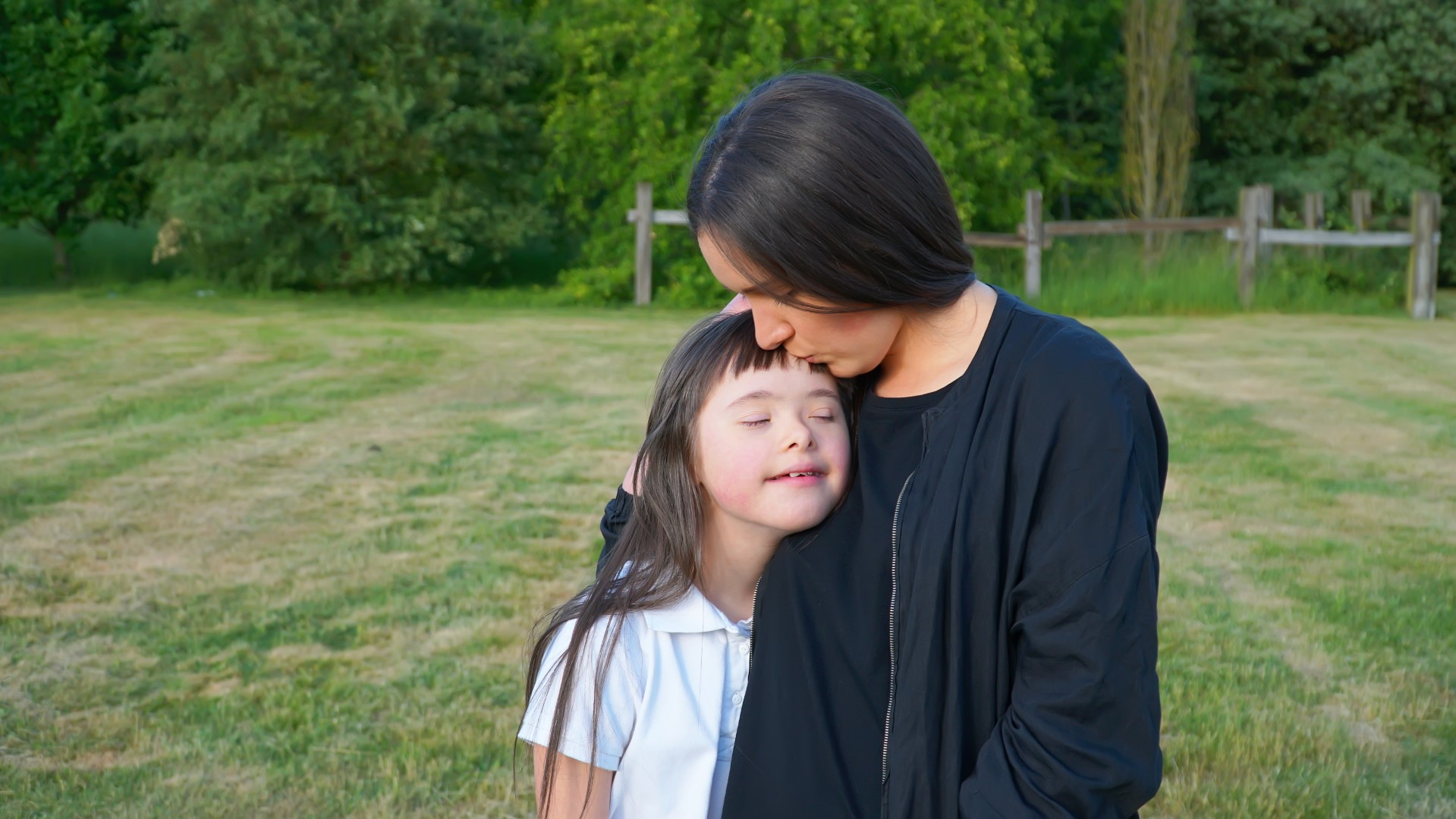 A carer hugs a child in a green outdoor space; the child has Down's syndrome.