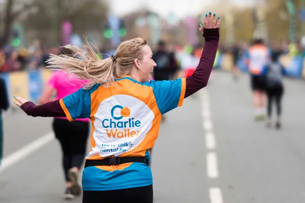 Female runner with blonde hair in a ponytail. She is taking part in a running race along a road. Her back is to the camera but she is looking off to the side, waving to spectators. She wears a running t-shirt with the Charlie Waller logo printed on. 