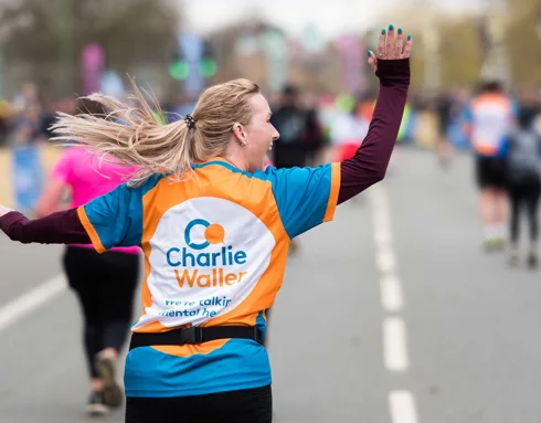 Female runner with blonde hair in a ponytail. She is taking part in a running race along a road. Her back is to the camera but she is looking off to the side, waving to spectators. She wears a running t-shirt with the Charlie Waller logo printed on.