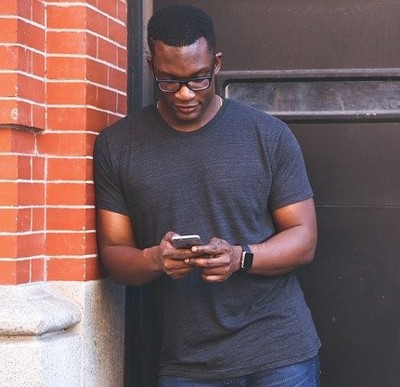 Black man leaning against wall typing on mobile phone